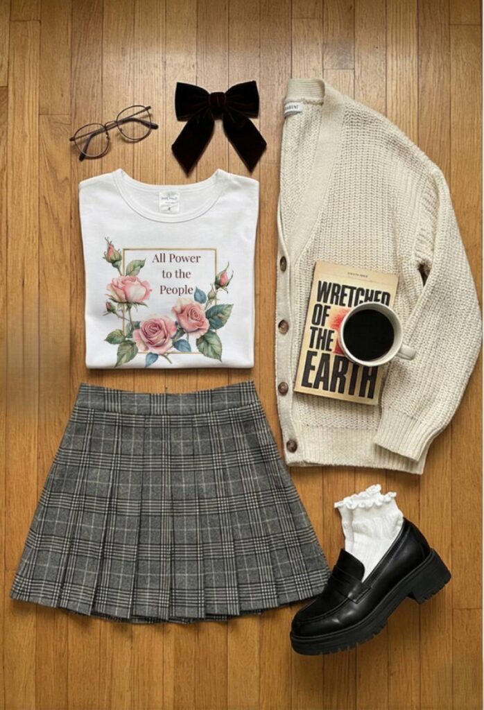 A Dark Academia outfit flat lay featuring a white 'All Power to the People' baby tee. The shirt is styled with a grey plaid pleated mini skirt, a cream chunky knit cardigan, and black platform loafers with white ruffle socks. Accessories include wire-rimmed glasses, a black velvet hair bow, and a copy of the book The Wretched of the Earth.